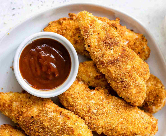 Golden, crispy breaded chicken tenders served on a white plate with a small bowl of barbecue sauce for dipping.