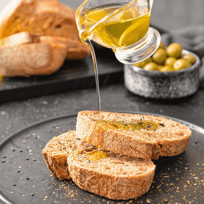 A stream of golden olive oil is being poured onto a slice of crusty bread resting on a white plate.