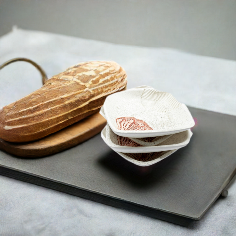 Four white plates are stacked on top of each other next to a white ceramic dipping plate with a garlic print design, all sitting on a black serving platter.  A fresh loaf of bread rests on a wooden cutting board in the background.