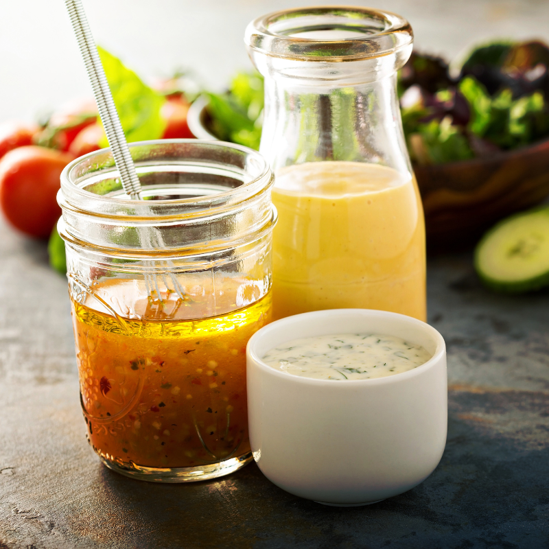 Several small glass containers filled with various vinaigrettes, oils, and dipping sauces are arranged together on a surface. A fresh, colorful salad is visible in the background.