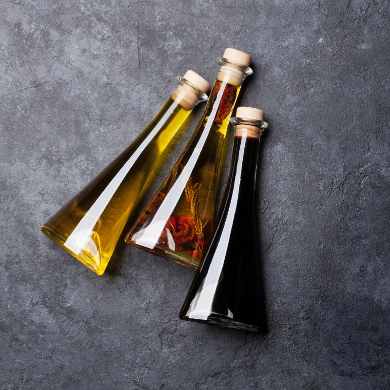 Three clear bottles of vinegar on a dark table. The vinegars are red, white, and brown.