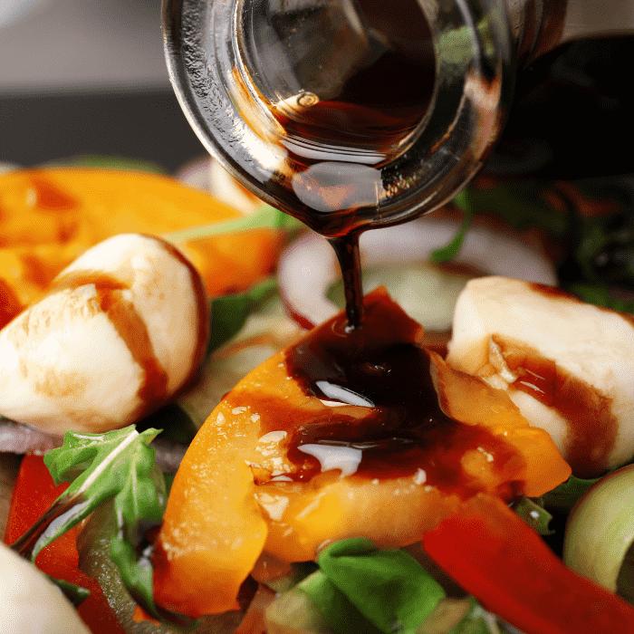 A thick stream of dark balsamic vinaigrette is being poured over a fresh salad filled with mozzarella cheese, sliced tomatoes, and leafy greens.