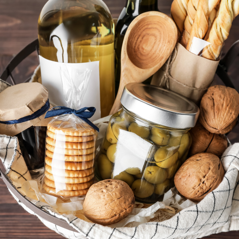 A gift basket filled with gourmet items like crackers, jam, olives, bread, walnuts, wine, and olive oil sits on a wooden table.