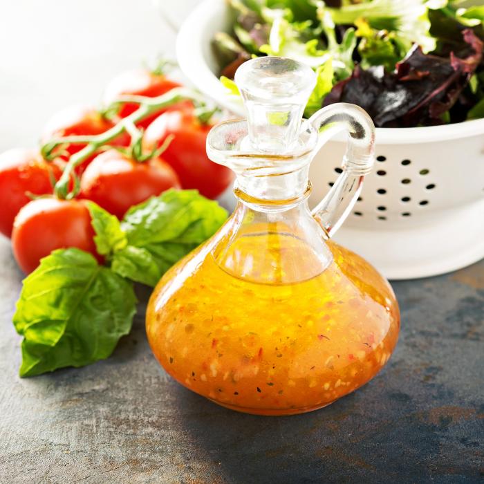 A clear glass bottle of white balsamic vinegar sits on a dark table. In the background, a colorful salad is visible in a colander and small vine oftomatoes.