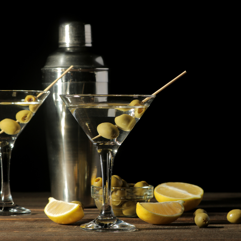 Two martini glasses filled with clear liquid and garnished with olives sit on a wooden table. Sliced lemons, whole olives, and a martini shaker are also arranged on the table.