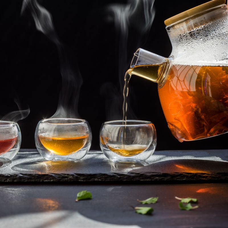 Red tea being poured from a glass teapot into a cup. Loose tea leaves are nearby.