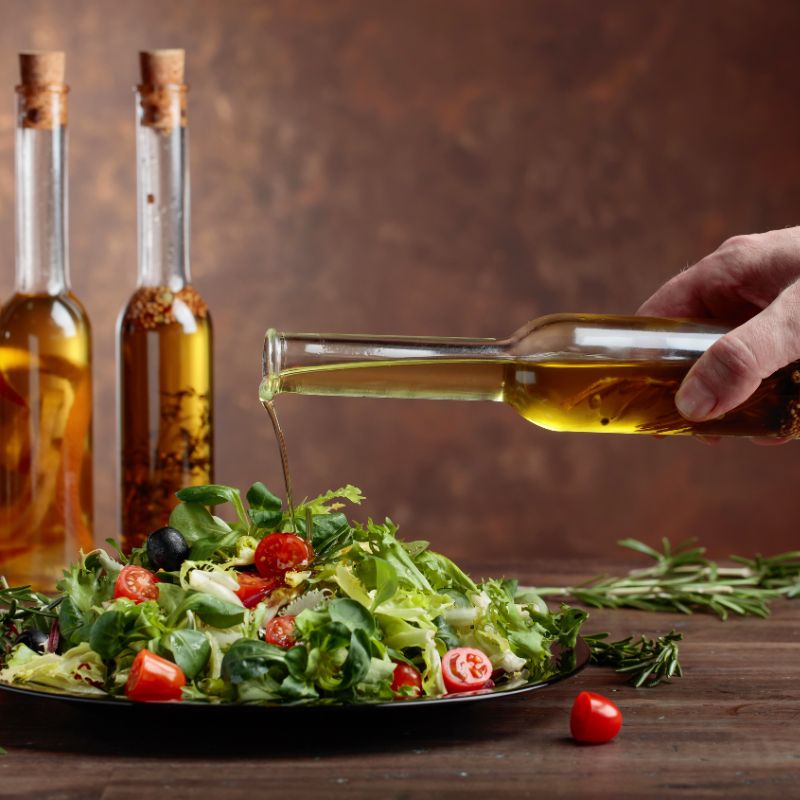 A person's hand pours infused olive oil from a clear bottle onto a vibrant salad on a wooden table. Several other bottles of infused oils are lined up in the background.