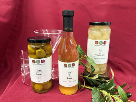 Buckeye Gourmet gift set with martini olive juice, cocktail onions, green olives, and martini glasses on red fabric.