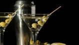 Two martini glasses with three skewered green olives in liquid on a dark brown table with a silver canister behind them. All with a black background behind. 