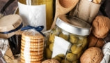 A close up shot of a beautiful basket with linen cloth holding a bottle of white wine, a jar of green olives, several walnuts, a package of crackers, a small jar of jam, and a bundle of breadsticks.